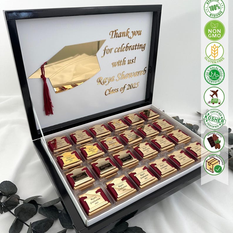 May include: A black gift box with a white interior, filled with chocolates. Each chocolate has a gold-colored plaque with text. The box lid features a gold graduation cap design and text that reads "Thank you for celebrating with us! Raya Shawareb Class of 2025."