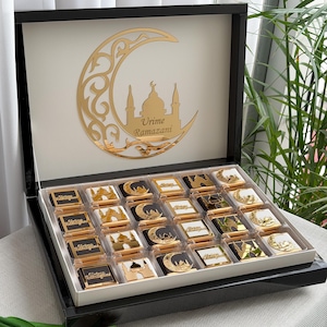 Könnte beinhalten: Eine schwarze Geschenkbox mit weißem Innenraum, gefüllt mit einzeln verpackten Pralinen. Der Deckel der Box zeigt ein goldenes Halbmond- und Moscheemotiv mit dem Text "Urime Ramazani". Jede Praline hat ein goldenes oder schwarzes Design.