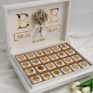 May include: A white gift box with gold lettering and a small floral arrangement. The box is open, displaying chocolates with gold foil wrappers. The lid features the names "BILAL" and "ELMA" and the date "02.14.2026".