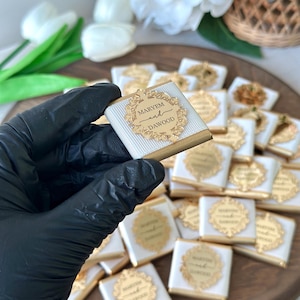 May include: Square chocolates with gold foil and white striped wrappers. Each chocolate has a gold emblem with the names "MARYEM and DAWOOD". The chocolates are arranged on a wooden tray, with some held in a gloved hand.