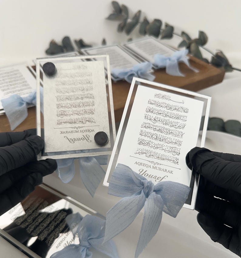 K&ouml;nnte beinhalten: Zwei silberne gerahmte Glasplaketten mit arabischer Kalligraphie und dem darunter gedruckten Namen "Yousef". Jede Plakette ist mit einem blauen Band gebunden.