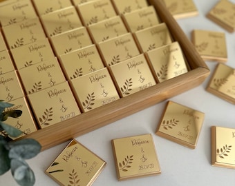 Puede incluir: Una caja de madera llena de barras de chocolate doradas. Cada barra está decorada con un diseño de hojas y el texto "Rebecca & Si 28.07.23".