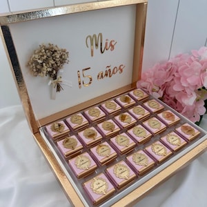 May include: A gold-framed box with a white interior, featuring the words "Mis 15 a&ntilde;os" in gold script. The box contains rows of pink-wrapped chocolates, each with a gold emblem. A small bouquet and pink flowers are also visible.