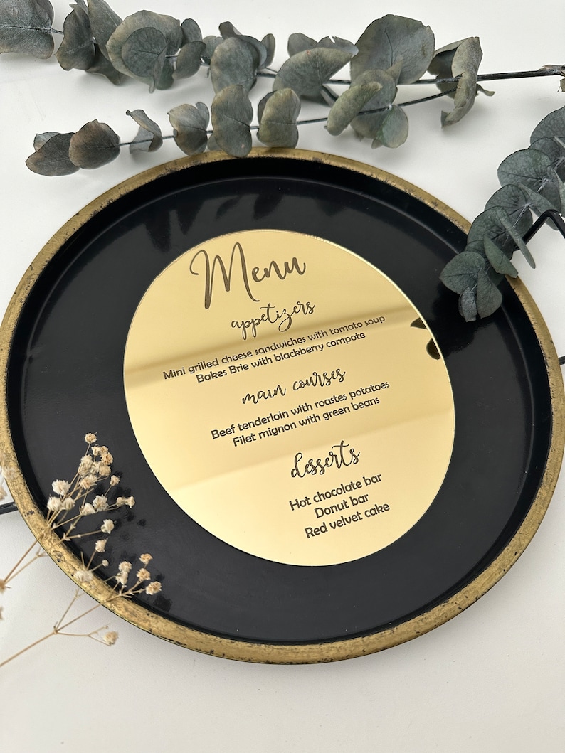 May include: A gold round menu sign with black text listing appetizers, main courses, and desserts. The sign is on a black plate with a gold rim.