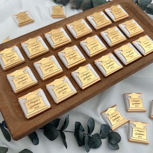 May include: A wooden tray holds numerous chocolate squares, each wrapped in white and gold foil. The gold foil features a graduation cap design and the text "Maryam's Graduation 2024". The chocolates are arranged on a white cloth with greenery.