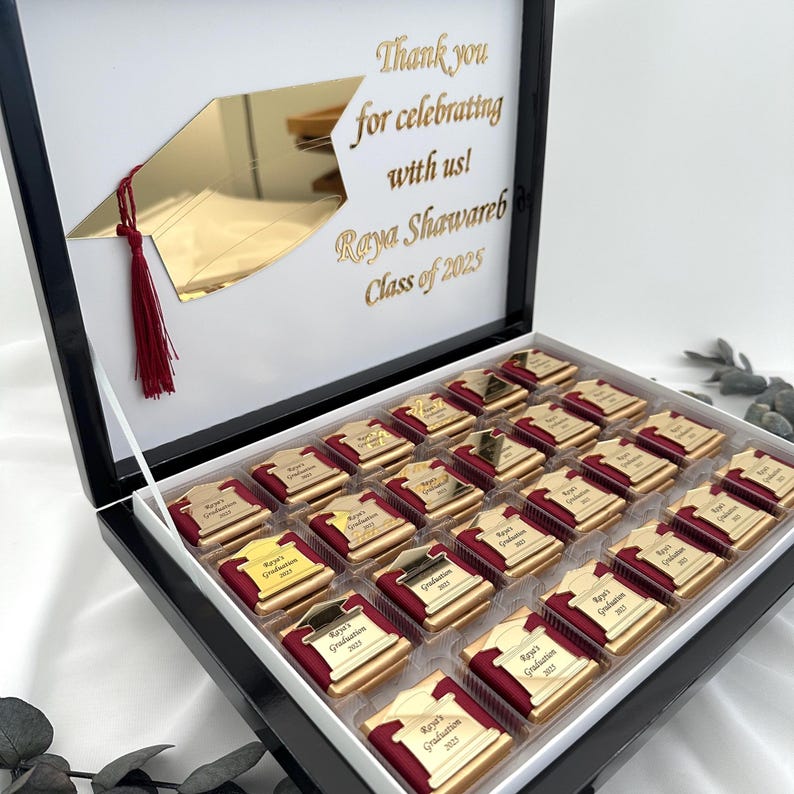 May include: A graduation gift box with a black frame and a white interior. The box contains chocolates with gold foil wrappers and burgundy accents. The lid features a gold graduation cap and the text "Thank you for celebrating with us! Raya Shawareb Class of 2025."