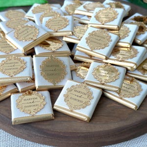 May include: An assortment of small, square chocolates wrapped in white and gold foil. Each chocolate is adorned with a gold label that reads "MARYEM and DAWOOD" in elegant script, set against a decorative border. The chocolates are arranged on a wooden tray.