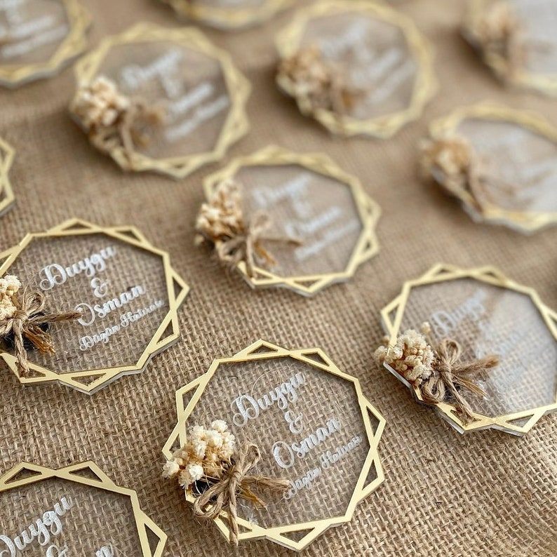 K&ouml;nnte beinhalten: Eine Sammlung von transparenten Acryl-Hochzeitsgeschenken mit goldenen geometrischen Rahmen. Jedes Geschenk enth&auml;lt einen kleinen Strau&szlig; getrockneter Blumen und die Namen "Duygu & Osman" mit dem Text "D&uuml;ğ&uuml;n Hatırası", was "Hochzeitsgedenken" bedeutet.