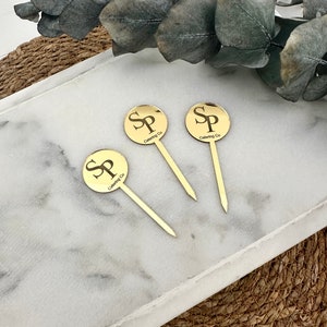 Könnte beinhalten: Drei goldfarbene Cocktailspieße aus Metall mit runden Köpfen. Jeder Kopf ist mit den Buchstaben "SP" und dem Text "Catering Co." graviert.