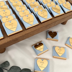 May include: Arrangement of small, wrapped chocolates on a wooden tray. Each chocolate is wrapped in light blue paper with gold trim and a gold heart-shaped label. The label has the text "Hacks & Millasay".
