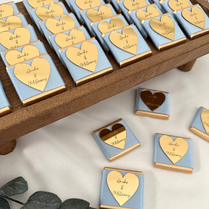 May include: Arrangement of small, wrapped chocolates on a wooden tray. Each chocolate is wrapped in light blue paper with gold trim and a gold heart-shaped label. The label has the text "Hacks & Millasay".