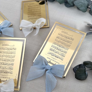 K&ouml;nnte beinhalten: Zwei goldene Rahmen mit arabischer Kalligraphie und einem blauen Band mit Schleife. Der Text auf den Rahmen lautet "AKIKAH MUBARAK" und "Jama".