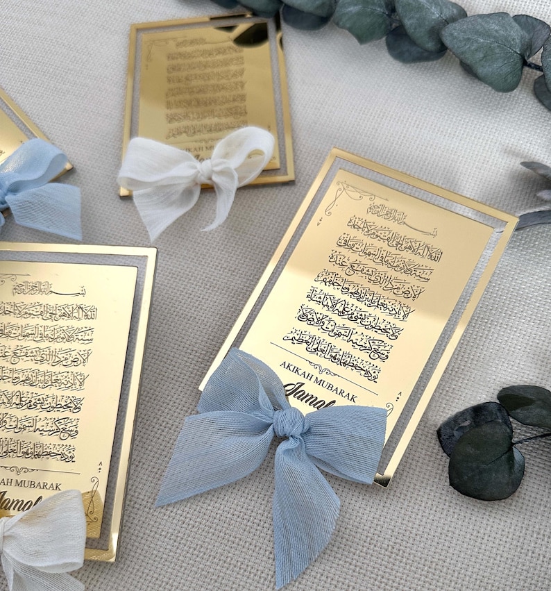 K&ouml;nnte beinhalten: Zwei goldene Rahmen mit arabischer Kalligraphie und einem blauen Band mit Schleife. Der Text auf den Rahmen lautet "AKIKAH MUBARAK" und "Jama".
