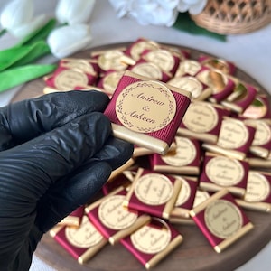 May include: A collection of small, wrapped chocolates with gold foil and burgundy ribbon. Each chocolate has a gold label with the names "Andrew & Ankeen" in a circular design. The chocolates are arranged on a wooden tray.