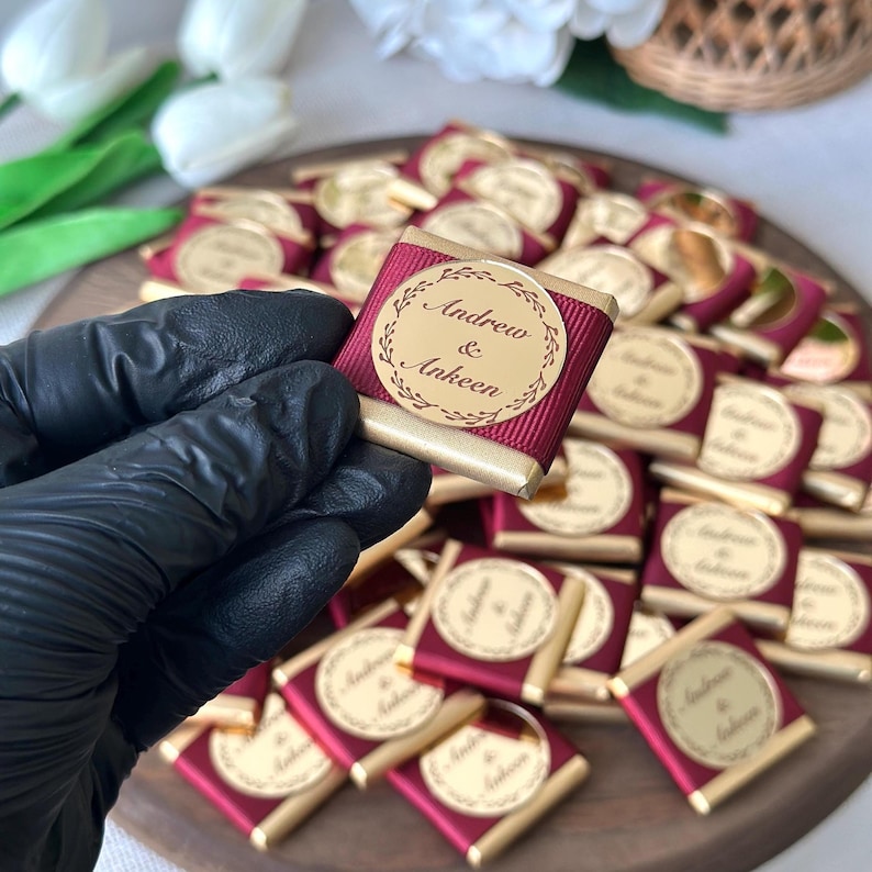 May include: A collection of small, wrapped chocolates with gold foil and burgundy ribbon. Each chocolate has a gold label with the names "Andrew & Ankeen" in a circular design. The chocolates are arranged on a wooden tray.