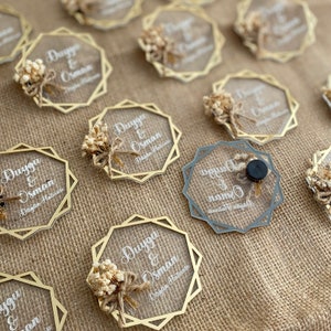 K&ouml;nnte beinhalten: Goldene und silberne geometrische Acryl-Hochzeitsgeschenke mit getrockneten Blumen und dem Text "Duygu & Osman" und "Duygu & Namik" auf einem Jute-Hintergrund.