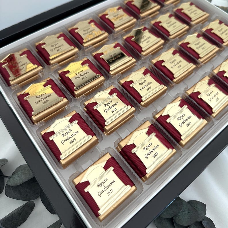 May include: A box of graduation chocolates. Each chocolate is wrapped in a burgundy ribbon and topped with a gold-colored plaque that reads "Raya's Graduation 2025". The chocolates are arranged in a black box.