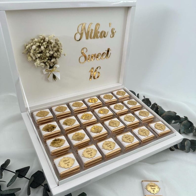 May include: A white gift box with a hinged lid, filled with individually wrapped chocolates. The box is decorated with gold lettering that reads "Nika's Sweet 16" and a small bouquet of white flowers. Each chocolate has a gold label.