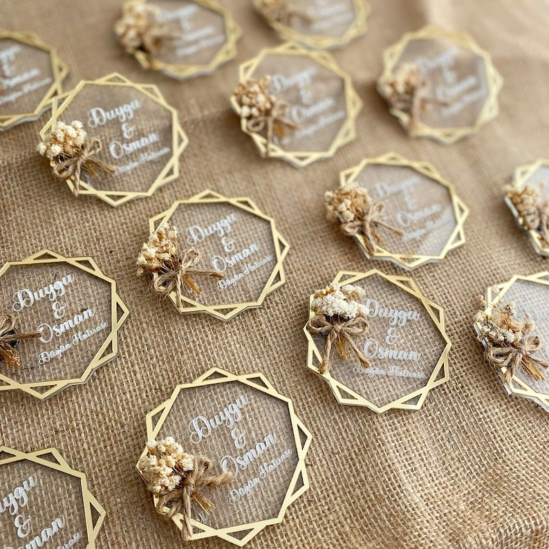 K&ouml;nnte beinhalten: Eine Sammlung von transparenten Acryl-Hochzeitsgeschenken mit goldenen geometrischen Rahmen. Jedes Geschenk zeigt die Namen "Duygu & Osman" und die Worte "D&uuml;ğ&uuml;n Hatırası" in wei&szlig;er Schrift. Die Geschenke sind mit kleinen Str&auml;u&szlig;en getrockneter wei&szlig;er Blumen verziert.