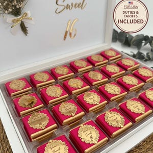 May include: A white box filled with individually wrapped chocolates. Each chocolate is wrapped in pink paper and topped with a gold-colored label that reads "Sweet 16". The box is decorated with gold lettering and a small floral arrangement.