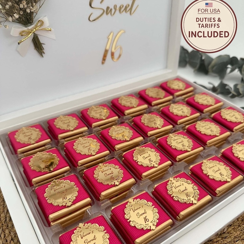 May include: A white box filled with individually wrapped chocolates. Each chocolate is wrapped in pink paper and topped with a gold-colored label that reads "Sweet 16". The box is decorated with gold lettering and a small floral arrangement.