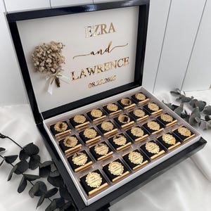 Peut inclure: Une boîte noire et blanche remplie de chocolats emballés individuellement. Le couvercle porte les noms "Ezra and Lawrence" en lettres dorées, ainsi que la date "08.25.2025". Un petit bouquet avec un ruban blanc est également présent.
