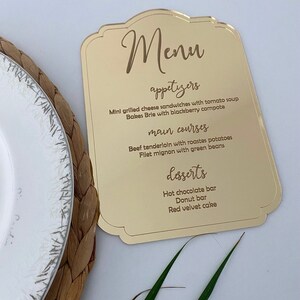 May include: Gold acrylic menu sign with a decorative border. The menu lists appetizers, main courses, and desserts. The text is in a cursive font.