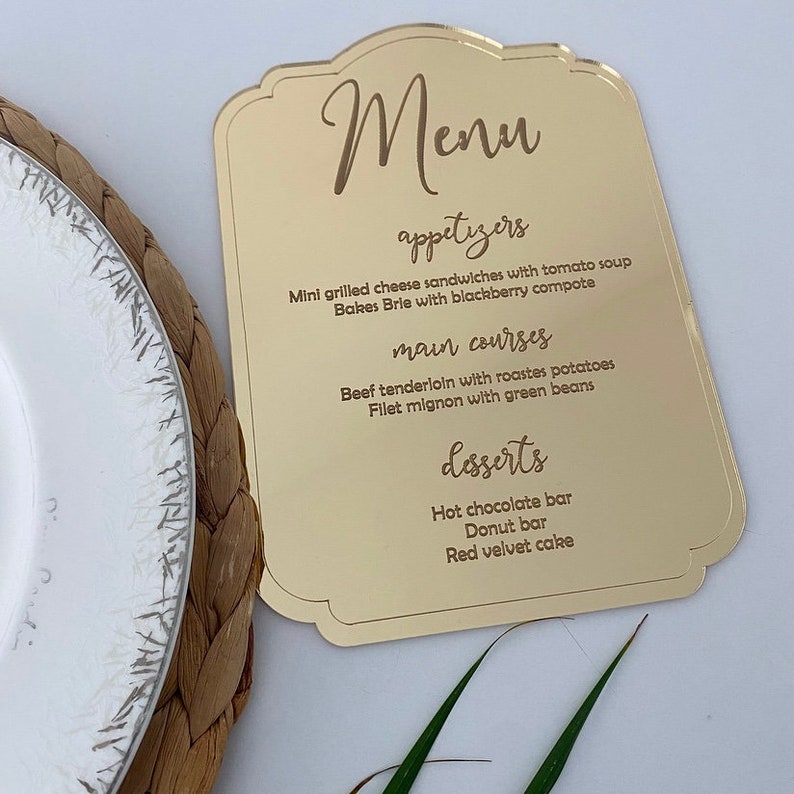 May include: Gold acrylic menu sign with a decorative border. The menu lists appetizers, main courses, and desserts. The text is in a cursive font.