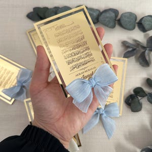 K&ouml;nnte beinhalten: Gold gerahmte Acryl-Plakette mit arabischer Kalligraphie und dem Text "AKIKAH MUBARAK Jamal" in goldenen Lettern. Eine hellblaue Schleife ist an der Unterseite der Plakette befestigt.