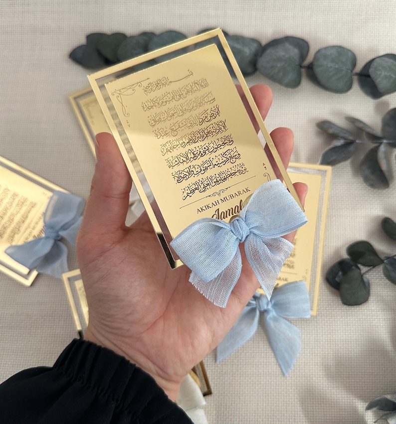 K&ouml;nnte beinhalten: Gold gerahmte Acryl-Plakette mit arabischer Kalligraphie und dem Text "AKIKAH MUBARAK Jamal" in goldenen Lettern. Eine hellblaue Schleife ist an der Unterseite der Plakette befestigt.