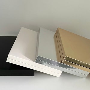 May include: A collection of four rectangular boxes in various colors: black, white, silver, and gold. The boxes are arranged in a row, with the black box on the left and the gold box on the right. The boxes have a glossy finish.