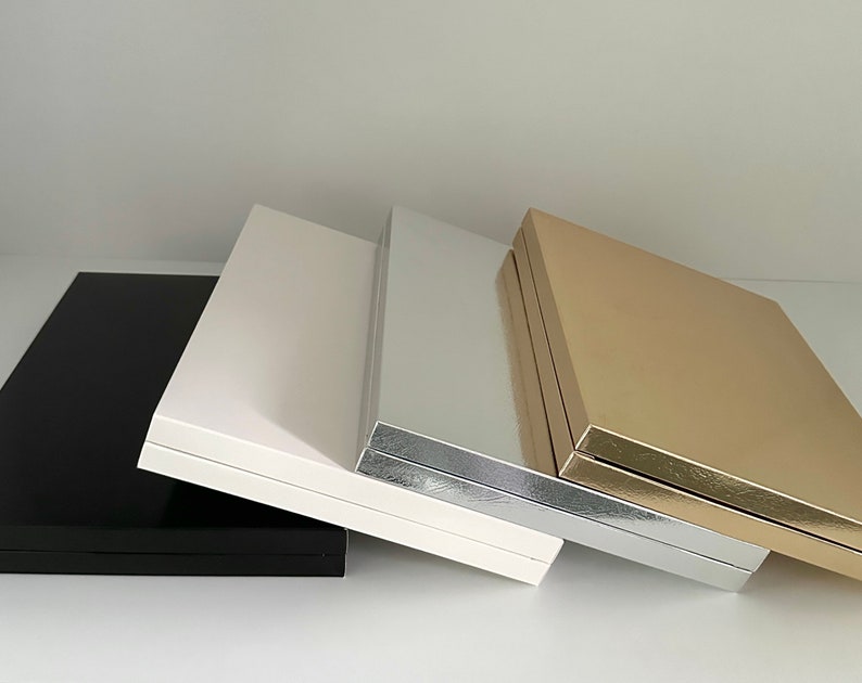 May include: A collection of four rectangular boxes in various colors: black, white, silver, and gold. The boxes are arranged in a row, with the black box on the left and the gold box on the right. The boxes have a glossy finish.