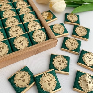 May include: A collection of personalized chocolates. Each chocolate is wrapped in dark green fabric and features a gold-colored emblem with the initials "N&B" and a heart. The chocolates are arranged in a wooden box and scattered on a white surface.