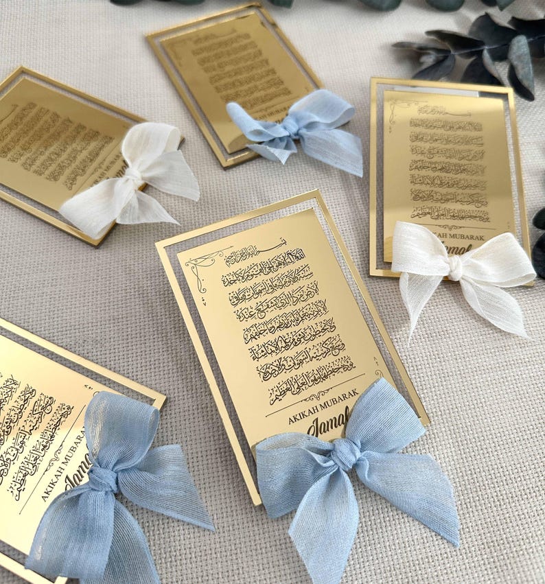 K&ouml;nnte beinhalten: Goldene Acryl-Plaketten mit arabischer Kalligraphie und dem Text "AKIKAH MUBARAK Jamal", verziert mit blauen und wei&szlig;en Schleifen.