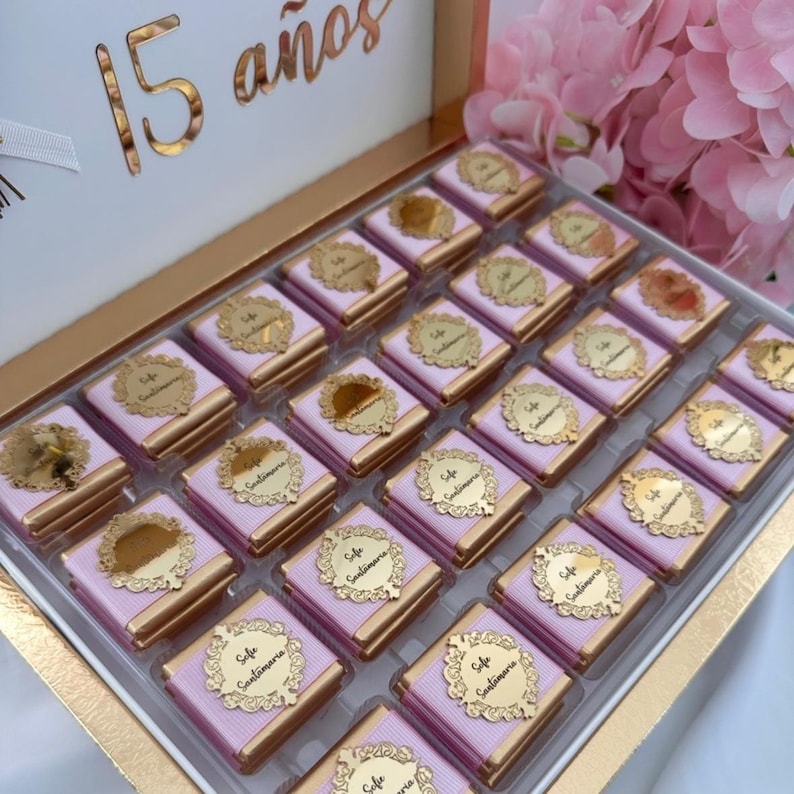 May include: A box of chocolates with pink and gold packaging. Each chocolate is wrapped in pink paper and topped with a gold label. The box is white with gold accents and the words "15 a&ntilde;os" in gold.