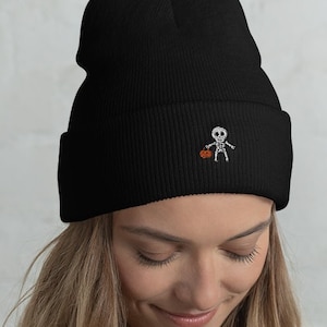 May include: Black beanie with a small embroidered skeleton holding a pumpkin. The beanie is made of a soft, ribbed fabric.