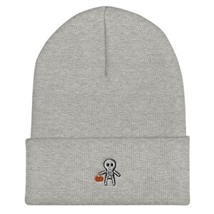 May include: Gray beanie with a small embroidered skeleton holding a pumpkin. The skeleton is wearing a white shirt and black pants.