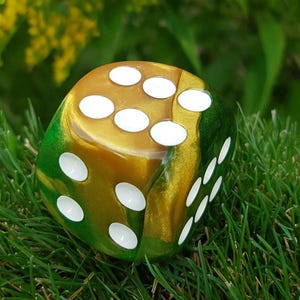 May include: A large, decorative die with a gold and green color scheme. The die has white dots and is resting on a bed of green grass. The background features yellow flowers and more green foliage.