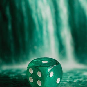 May include: A close-up of a translucent green die with white dots, showing the numbers 1, 2, 3, and 5. The die is set against a blurred background of green and white, resembling a waterfall. The die is a gaming accessory.