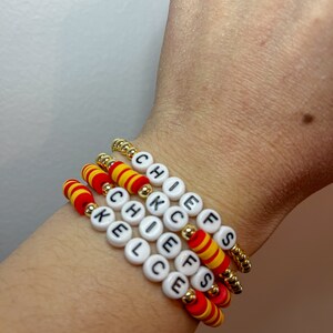 May include: Four beaded bracelets spelling "CHIEFS" and "KELCE" in white letter beads. The bracelets feature red and yellow striped beads and gold-colored spacer beads. The bracelets are stacked on a wrist.