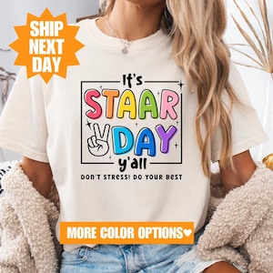 May include: White t-shirt with a colorful graphic that says "It's STAAR DAY y'all DON'T STRESS! DO YOUR BEST".