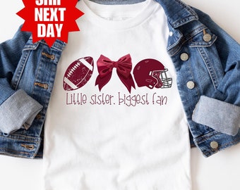 Puede incluir: Camiseta blanca con un gráfico de fútbol americano, un lazo y un casco marrones. El texto "Little sister. Biggest fan." está impreso debajo del gráfico.