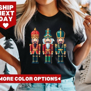 May include: A black t-shirt featuring a graphic of three nutcracker soldiers. The nutcrackers are dressed in red, green and blue uniforms. The shirt has the text "MORE COLOR OPTIONS" printed at the bottom.