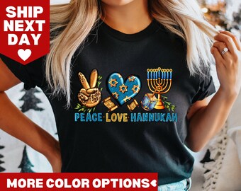 Könnte beinhalten: Schwarzes T-Shirt mit einem Peace-Zeichen, einem Herzen, einem Chanukka-Leuchter und einem Dreidel. Der Text "Peace Love Hanukkah" ist unter dem Motiv gedruckt.