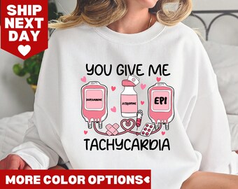 Op de afbeelding: Wit sweatshirt met een roze afbeelding van een hart, infuuszakken en medische benodigdheden. De tekst luidt "You give me tachycardia".