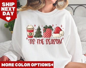 May include: White sweatshirt with a Christmas-themed design featuring a cup of coffee, a Christmas tree, a gift box, and a Santa hat. The text "'Tis the Season" is printed below the design.