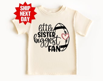 Puede incluir: Una camiseta blanca con un gráfico negro que dice "little sister biggest fan" con un corazón rojo y un gráfico de fútbol.