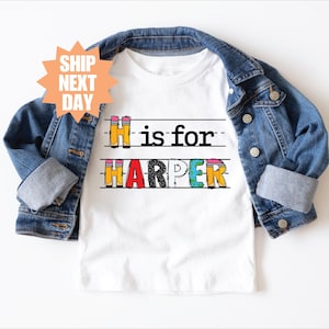 May include: A white short-sleeved t-shirt featuring the text H is for HARPER in a colorful pencil-themed font, layered under a blue denim jacket with metal buttons. The design includes horizontal lines resembling lined school paper.