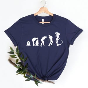 May include: A navy blue t-shirt with a white graphic of the evolution of humans to an alien. The graphic shows a series of silhouettes starting with a single-celled organism, then a caveman, then a modern human, and finally an alien.