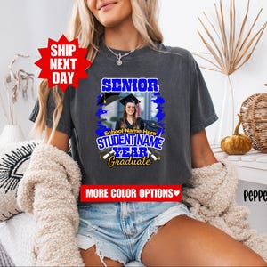 May include: A gray t-shirt with a blue and yellow graphic design that says "Senior" and "Graduate". The graphic also includes a photo of a person wearing a graduation cap and gown. The text "School Name Here" and "Student Name" are also included in the graphic.